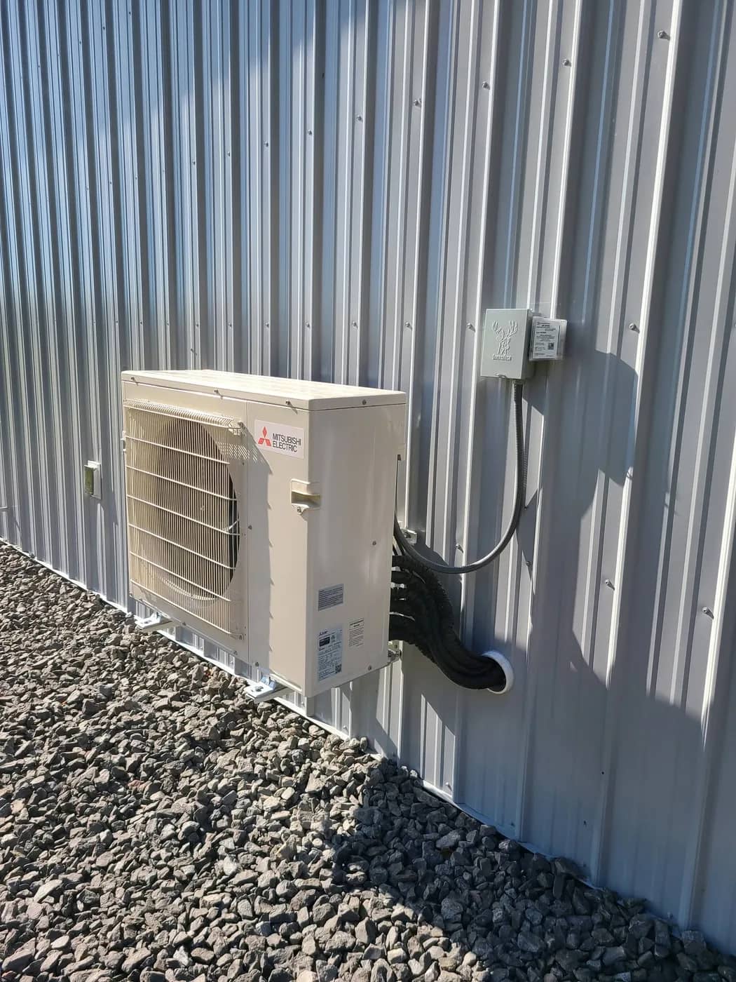 Mitsubishi Electric outdoor heat pump condenser mounted on brackets beside a metal-sided building, with line-set and disconnect box