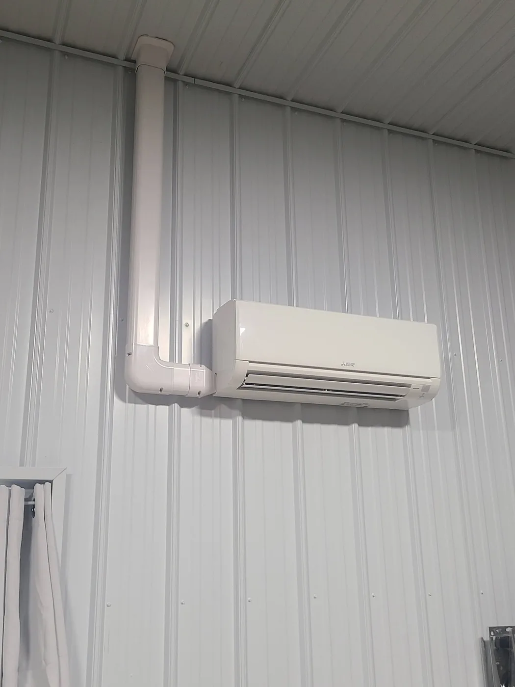 Mitsubishi ductless mini-split wall head installed high on a metal shop wall with white line-set cover running to the ceiling